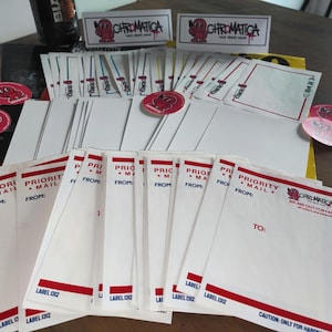 May include: An assortment of white and red "Chromatica" branded stickers, labels, and envelopes. The envelopes are labeled "Priority Mail" and have a red stripe. Some stickers are round with a cartoon character.