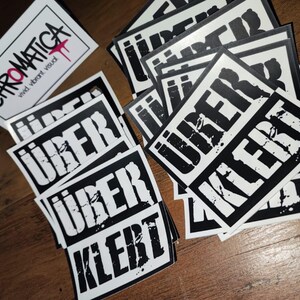 Op de afbeelding: Een stapel zwart-wit stickers met het woord "UBER" en "KLEBT" erop gedrukt. De stickers liggen op een houten oppervlak.