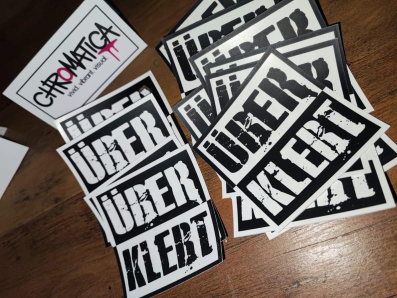 Op de afbeelding: Een stapel zwart-wit stickers met het woord "UBER" en "KLEBT" erop gedrukt. De stickers liggen op een houten oppervlak.