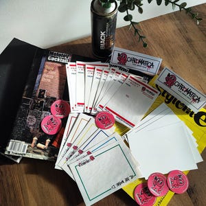 Puede incluir: Un surtido de materiales de arte y promocionales, que incluyen una lata de pintura en aerosol negra, pegatinas, botones, sobres y una revista. Los artículos están dispuestos sobre una superficie de madera, con una planta en una lata reutilizada en el fondo.