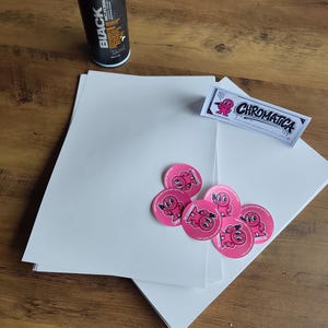 Könnte beinhalten: Eine schwarze Sprühdose, ein rechteckiger Aufkleber mit dem Text CHROMATICA und einer rosa Zeichentrickfigur sowie mehrere runde rosa Aufkleber mit derselben Figur, angeordnet auf einem Stapel weißer, rechteckiger Papierbögen.