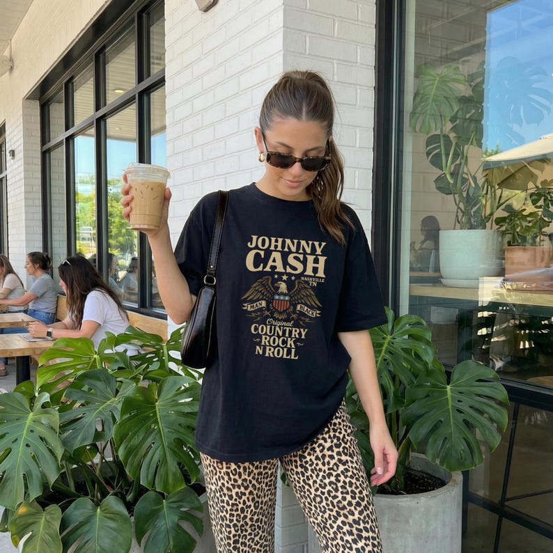 May include: Black t-shirt featuring the text "JOHNNY CASH" above an eagle graphic. The shirt also includes the words "Original Country Rock N Roll" and is paired with leopard print pants. The person is holding a beverage.