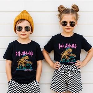 May include: Two children wearing black t-shirts with a Def Leppard graphic featuring a leopard and the word "ANIMAL". The shirts have a pink and teal design. One child wears a yellow beanie and the other has their hair in buns. Both wear sunglasses and checkered shorts.