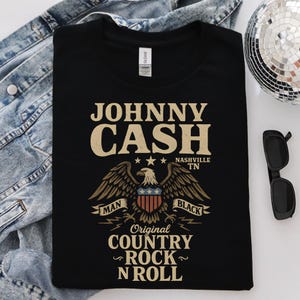May include: Black t-shirt featuring the text "JOHNNY CASH" above an eagle graphic with the words "MAN" and "BLACK" on banners. The shirt also includes "Original COUNTRY ROCK N ROLL" and "NASHVILLE TN".