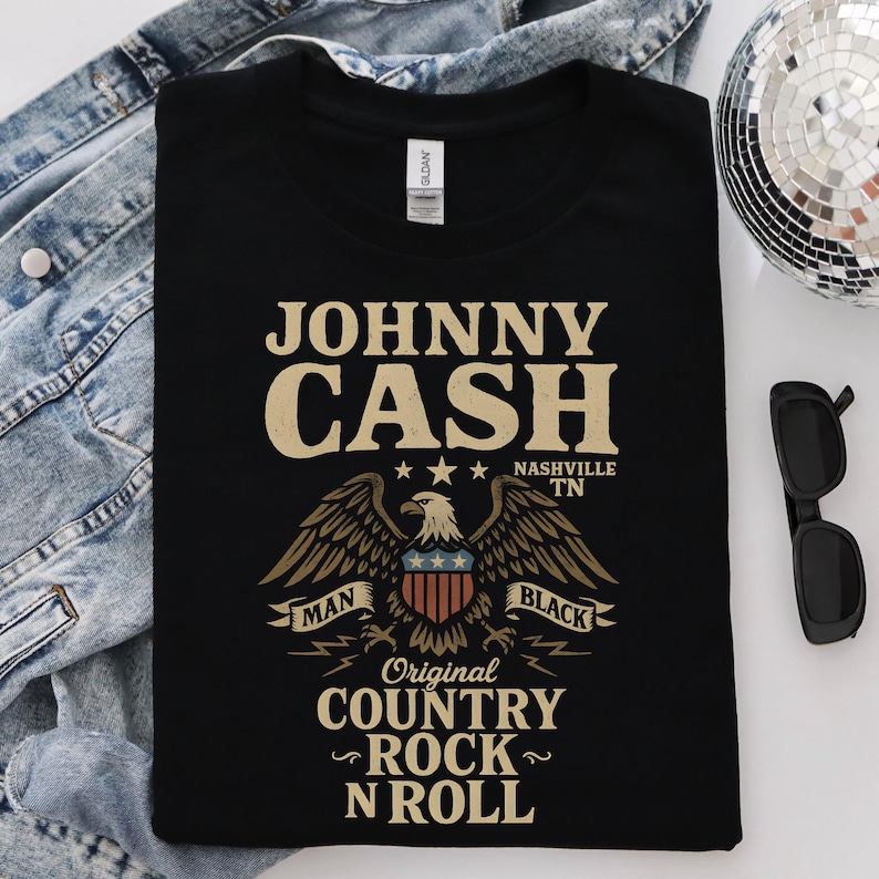 May include: Black t-shirt featuring the text "JOHNNY CASH" above an eagle graphic with the words "MAN" and "BLACK" on banners. The shirt also includes "Original COUNTRY ROCK N ROLL" and "NASHVILLE TN".