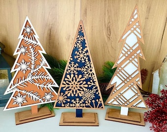 May include: Three wooden Christmas tree decorations with different designs. The trees are white with brown accents and feature snowflake patterns. The trees are on wooden stands.