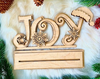 Puede incluir: Corte de madera de la palabra "ALEGRÍA" con un gorro de Papá Noel en la "A". El corte está decorado con copos de nieve y tiene un espacio en blanco para un mensaje.