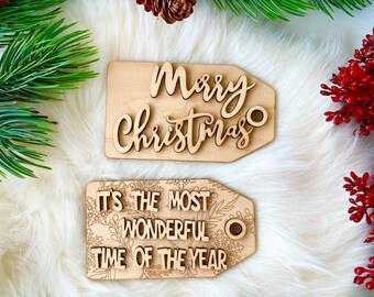 May include: Two wooden Christmas gift tags with a white background. The top tag says "Merry Christmas" in a cursive font. The bottom tag says "It's the most wonderful time of the year" with a floral design.
