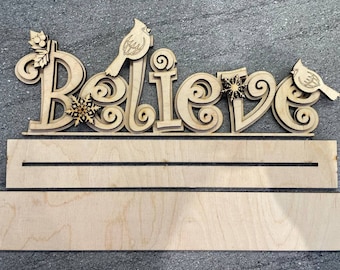 Puede incluir: Un letrero de madera con la palabra "Believe" en una fuente decorativa. El letrero presenta dos cardenales y copos de nieve. El letrero está sin terminar y listo para ser pintado o teñido.