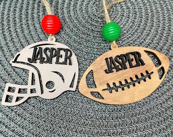 May include: Two wooden ornaments with the name "Jasper" on them. One ornament is shaped like a football and the other is shaped like a football helmet. Both ornaments are painted brown and have black details.