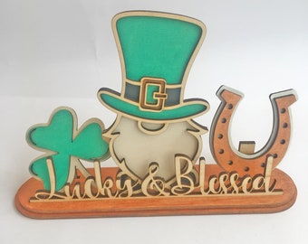 May include: Wooden cutout of a leprechaun with a green hat, a shamrock, and a horseshoe. The cutout is on a wooden base with the words "Lucky & Blessed" written on it.