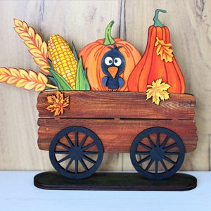 May include: A wooden wagon filled with fall harvest items, including pumpkins, corn, wheat, and a black bird. The wagon has black wheels and is on a wooden base.