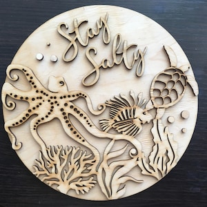 May include: A wooden round sign with the text "Stay Salty" in a cursive font. The sign features a cutout design of an octopus, a fish, a turtle, and seaweed.