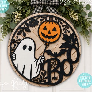 May include: A circular wooden wall sign featuring a white ghost holding an orange jack-o'-lantern balloon, a black bat, and the word BOO in black lettering. The background has a light wood chevron pattern, and the sign is accented with black leaves.