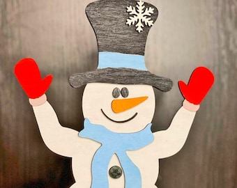 May include: A wooden snowman figurine with a black top hat, blue scarf, and red mittens. The snowman has a white body, orange nose, and black eyes. The figurine is decorated with snowflakes and has a pink base.