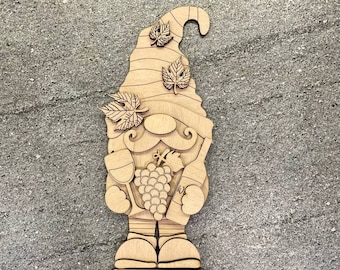 May include: Wooden gnome figurine holding a wine glass and bottle of wine. The gnome is wearing a hat with leaves and has a beard. The gnome is standing on a wooden base.