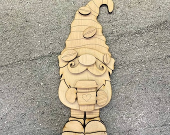 May include: Wooden gnome figurine holding a coffee cup with a heart on it. The gnome has a brown hat with coffee beans on it. The gnome is standing on a wooden base. There are two additional wooden bases.