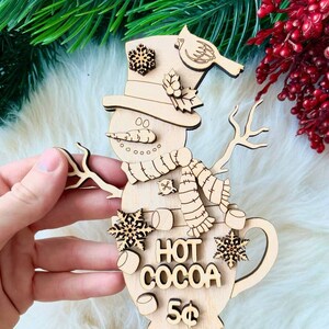May include: Wooden snowman ornament shaped like a mug with the words "Hot Cocoa 5¢" on it. The snowman has a carrot nose, a scarf, and a hat with a snowflake on top. There are also snowflakes and marshmallows on the mug.