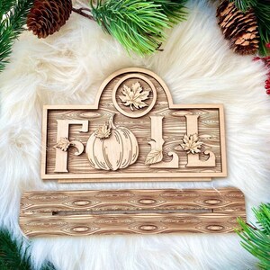 Op de afbeelding: Een houten bord met het woord "FALL" in grote letters, versierd met gesneden pompoenen en bladeren. Het bord heeft een houtnerfpatroon en is perfect voor herfstdecoratie.