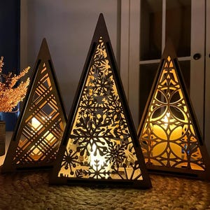 May include: Three wooden, triangular candle holders with intricate cut-out designs. The candle holders are illuminated from within, casting a warm glow. The designs include snowflakes, geometric patterns, and floral motifs. The candle holders are on a woven mat.