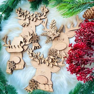 May include: Three wooden reindeer ornaments with intricate details. Each reindeer has a different design, including a floral wreath, a snowflake, and a garland of lights. The ornaments are on a white fluffy background.