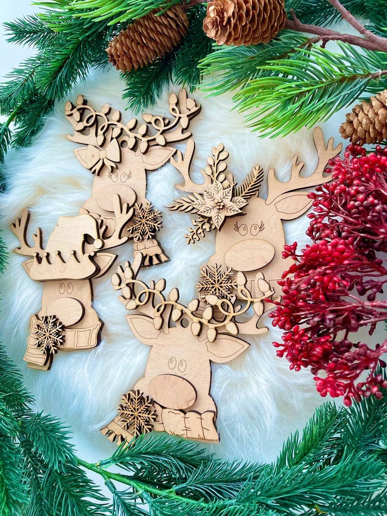 May include: Three wooden reindeer ornaments with intricate details. Each reindeer has a different design, including a floral wreath, a snowflake, and a garland of lights. The ornaments are on a white fluffy background.
