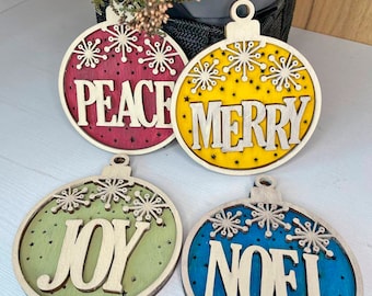 May include: Four wooden Christmas ornaments with a snowflake design. Each ornament is painted a different color and has a word on it: Peace, Merry, Joy, and Noel.