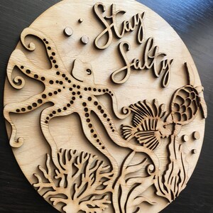 Op de afbeelding: Een houten bord met een uitgesneden ontwerp van een octopus, een vis en koraal. Het bord heeft de tekst "Stay Salty".
