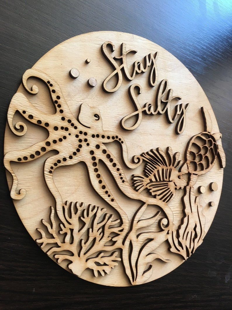 Op de afbeelding: Een houten bord met een uitgesneden ontwerp van een octopus, een vis en koraal. Het bord heeft de tekst "Stay Salty".