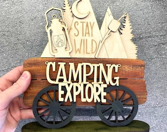 May include: Wooden sign with a wagon wheel design. The sign says "Camping Explore" and has a smaller sign above it that says "Stay Wild". The smaller sign has a crescent moon, mountains, a campfire, and a lantern.