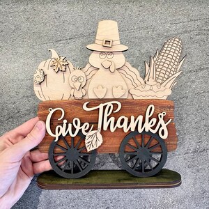 K&ouml;nnte beinhalten: Ein h&ouml;lzernes Thanksgiving-Schild mit einem Truthahn, einem K&uuml;rbis, Mais und einem Kuchen. Das Schild sagt "Give Thanks" und hat R&auml;der.