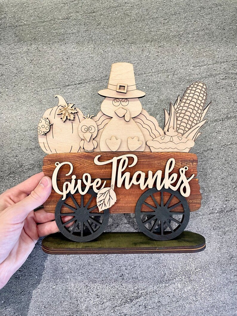 K&ouml;nnte beinhalten: Ein h&ouml;lzernes Thanksgiving-Schild mit einem Truthahn, einem K&uuml;rbis, Mais und einem Kuchen. Das Schild sagt "Give Thanks" und hat R&auml;der.