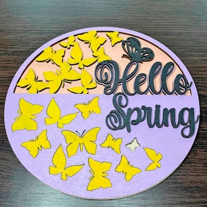 May include: A round wooden sign with a purple and pink background. The sign says "Hello Spring" in black lettering. The sign is decorated with yellow butterflies.