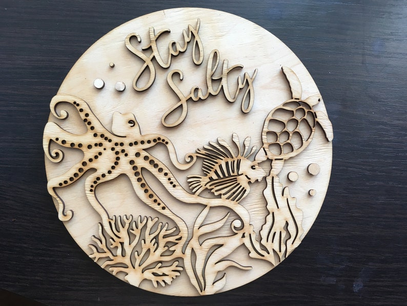 Op de afbeelding: Een rond houten bord met de tekst "Stay Salty" in een cursief schrift. Het bord heeft een uitgesneden ontwerp van een octopus, een vis, een schildpad en zeewier.