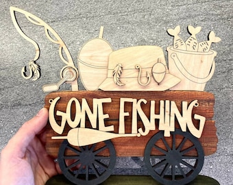 May include: Wooden sign with the words "Gone Fishing" on a wagon wheel design. The sign is decorated with fishing-themed items such as a fishing rod, a hat, a bucket, and fish.