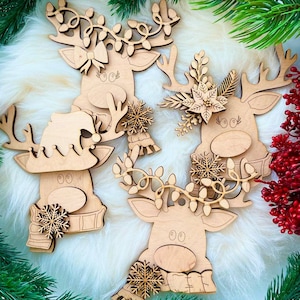 May include: Three wooden reindeer ornaments with different decorations. The reindeer on the left has a snowflake and a scarf, the reindeer in the middle has a floral design, and the reindeer on the right has a garland of lights.