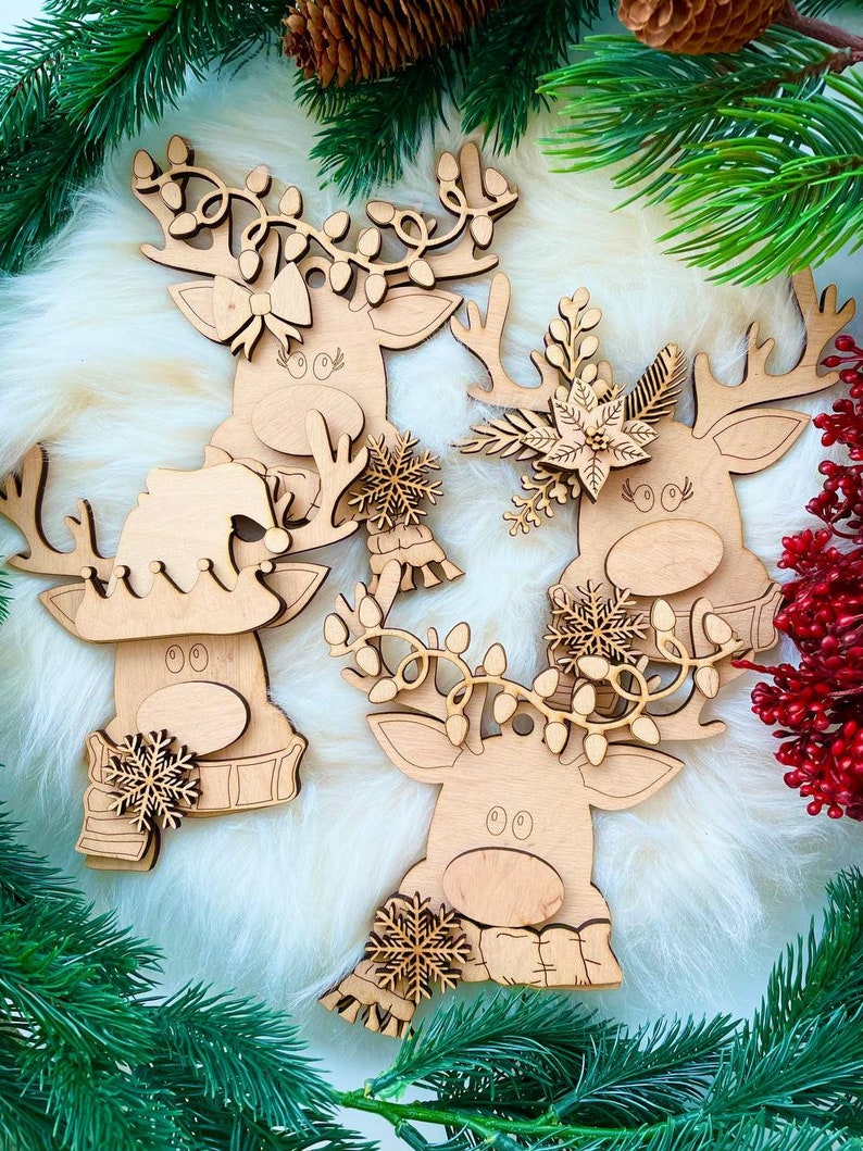 May include: Three wooden reindeer ornaments with different decorations. The reindeer on the left has a snowflake and a scarf, the reindeer in the middle has a floral design, and the reindeer on the right has a garland of lights.