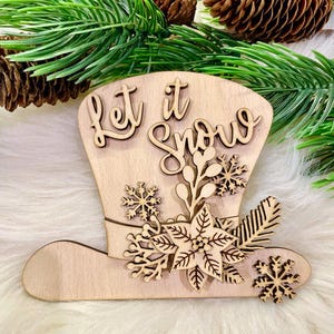 Puede incluir: Adorno navideño de madera con forma de sombrero con las palabras "Let it Snow" en la parte superior. El sombrero está decorado con copos de nieve, poinsettias y vegetación.