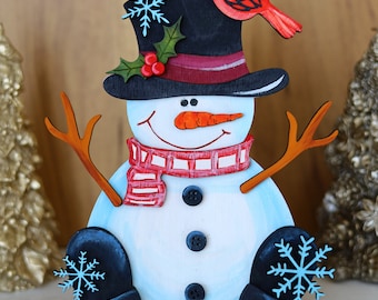 Op de afbeelding: Een houten sneeuwpopfiguur met een zwarte hoge hoed, een rood-wit gestreepte sjaal en een wortelneus. De sneeuwpop heeft twee zwarte knopen en een blauwe sneeuwvlok op elke hand. Een rode kardinaal zit op de hoed.