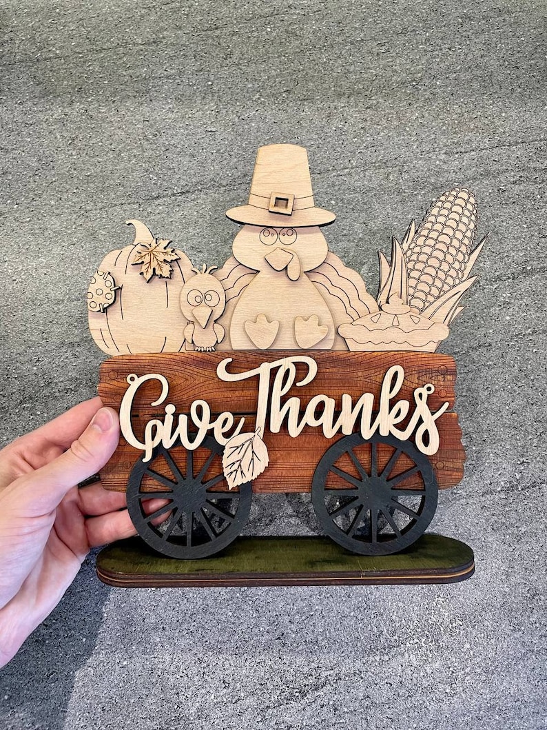 K&ouml;nnte beinhalten: Ein h&ouml;lzernes Thanksgiving-Schild mit einem Truthahn, einem K&uuml;rbis, Mais und einem Kuchen in einem Wagen. Das Schild sagt "Give Thanks".