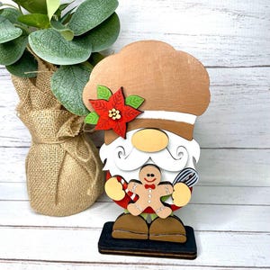 Könnte beinhalten: Eine hölzerne Weihnachts-Gnom-Figur mit einem braunen Hut, weißem Bart und rot-weißer Kleidung. Der Gnom hält einen Lebkuchenmann und einen Schneebesen. Der Gnom steht auf einem schwarzen Sockel.