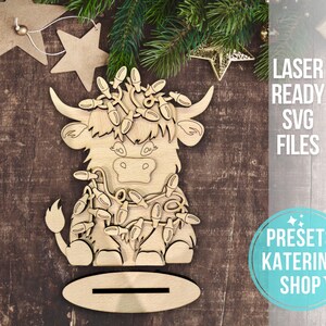 May include: A wooden cutout of a Highland cow wearing a string of Christmas lights. The cutout is designed to stand upright and has a slot in the base for a stand.