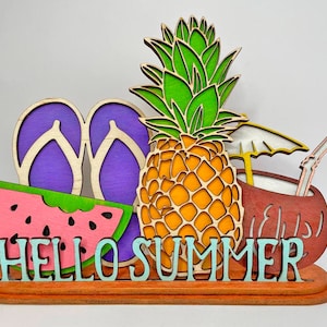 Op de afbeelding: Houten uitsnijding van een ananas, een watermeloen, slippers en een kokosnoot met een rietje en een paraplu. De uitsnijding is op een houten basis met de tekst "HELLO SUMMER" in blauw.