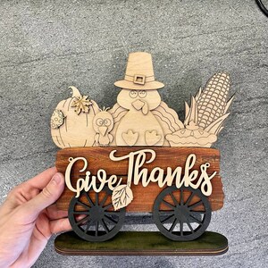K&ouml;nnte beinhalten: Ein h&ouml;lzernes Thanksgiving-Schild mit einem Truthahn, einem K&uuml;rbis, Mais und einer Torte. Das Schild lautet "Give Thanks" und hat R&auml;der.