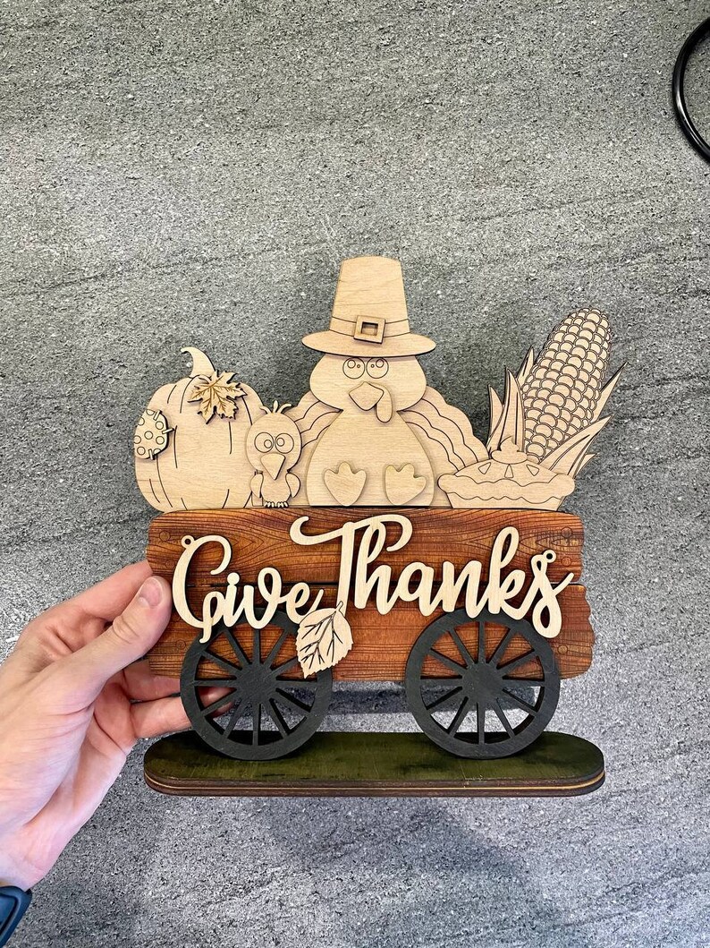 K&ouml;nnte beinhalten: Ein h&ouml;lzernes Thanksgiving-Schild mit einem Truthahn, einem K&uuml;rbis, Mais und einer Torte. Das Schild lautet "Give Thanks" und hat R&auml;der.