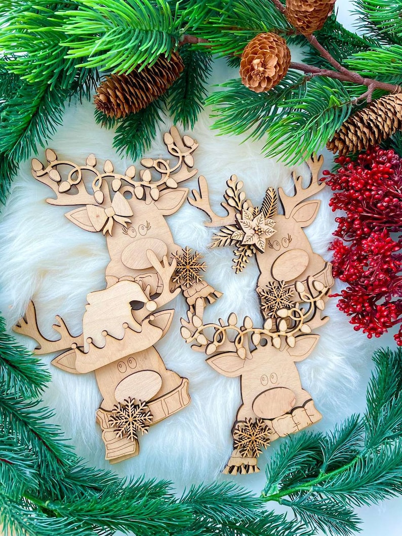 May include: Three wooden reindeer ornaments with different Christmas decorations. The reindeer are painted in a natural wood color and have white accents. The ornaments are surrounded by green pine branches and red berries.