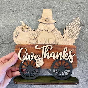 K&ouml;nnte beinhalten: Ein h&ouml;lzernes Thanksgiving-Schild mit einem Truthahn, einem K&uuml;rbis, Mais und den Worten "Give Thanks" auf einem Wagen.