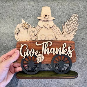 K&ouml;nnte beinhalten: Ein h&ouml;lzernes Thanksgiving-Schild mit einem Truthahn, einem K&uuml;rbis, Mais und einer Torte. Das Schild lautet "Give Thanks" und hat R&auml;der.