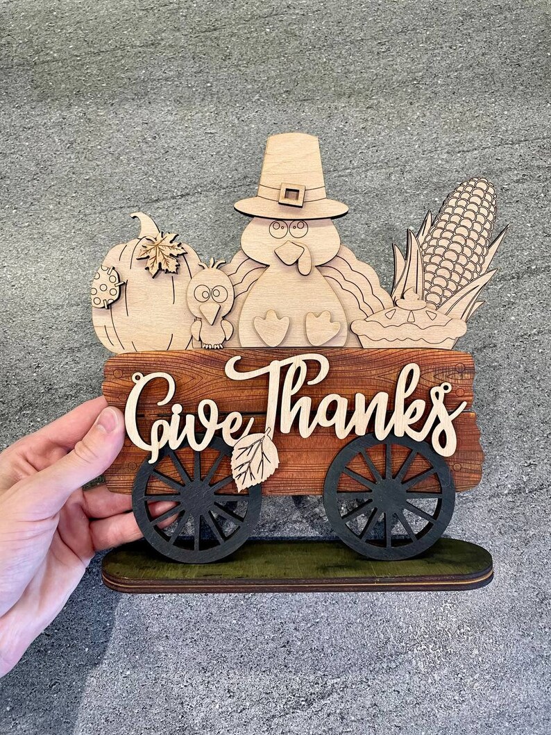 K&ouml;nnte beinhalten: Ein h&ouml;lzernes Thanksgiving-Schild mit einem Truthahn, einem K&uuml;rbis, Mais und einer Torte. Das Schild lautet "Give Thanks" und hat R&auml;der.