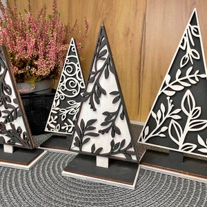 Peut inclure: Quatre décorations d'arbre de Noël en bois noir et blanc avec des motifs découpés complexes. Les arbres sont posés sur des bases noires et sont disposés sur une surface texturée grise.